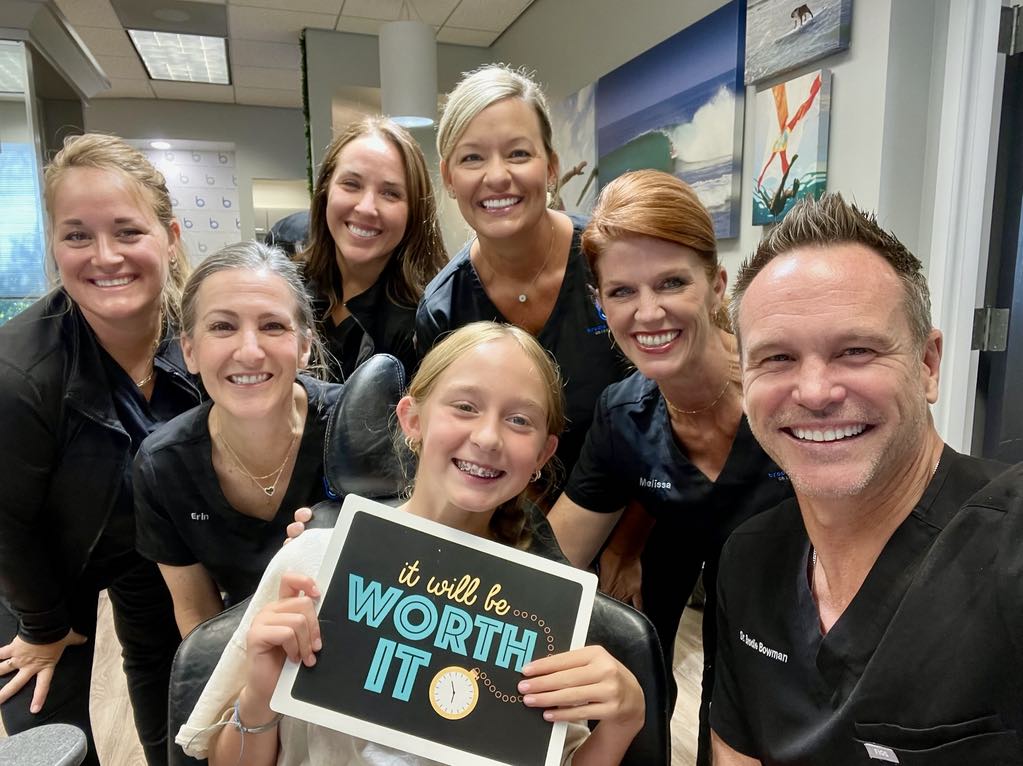 A young Brodie Bowman Orthodontics patient in braces holding an 'It will be WORTH IT' sign in the treatment chair, surrounded by Dr. Bowman and the team at the Fort Walton Beach office