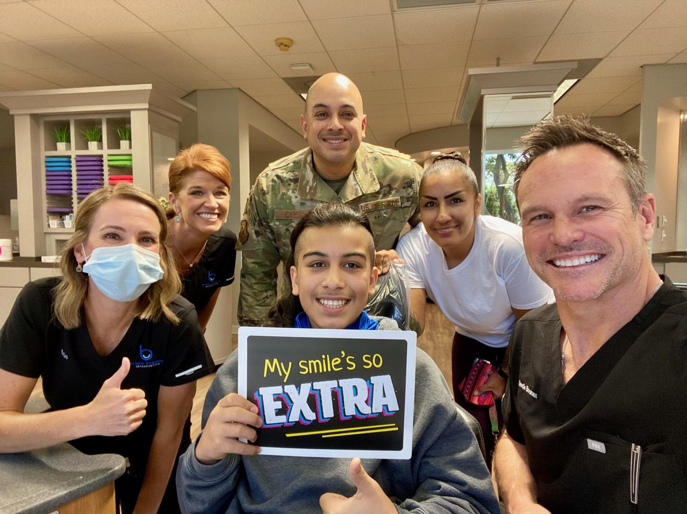 A teen Brodie Bowman Orthodontics patient in the treatment chair holding a My Smile's So Extra sign, with her Air Force dad, mom, Dr. Bowman and the team