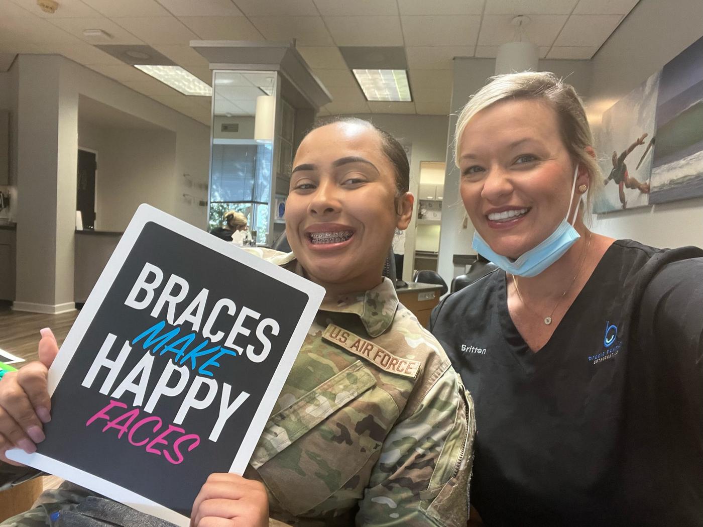 An Air Force patient in braces holding a Braces Make Happy Faces sign with assistant Britten at Brodie Bowman Orthodontics